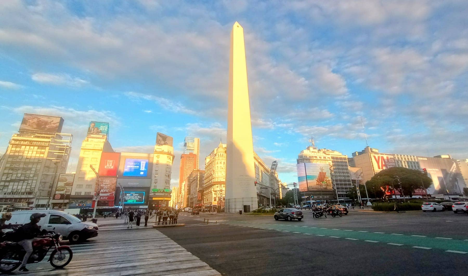 The Obelisco of Buenos Aires in warm evening light, with billboards, avenue traffic and buildings around it
