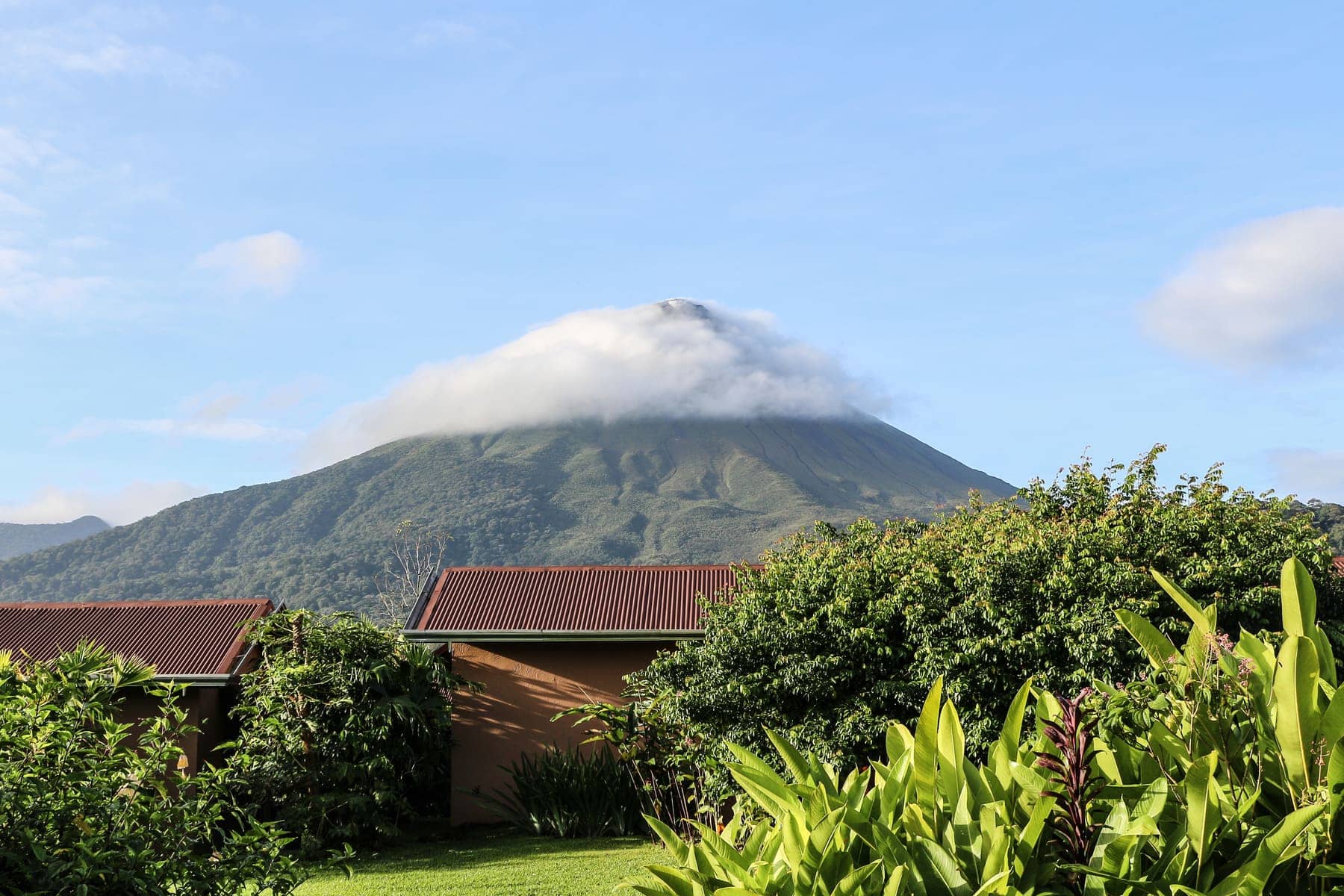 The cloud-capped cone of Arenal Volcano rising above tropical vegetation and low red-roofed buildings