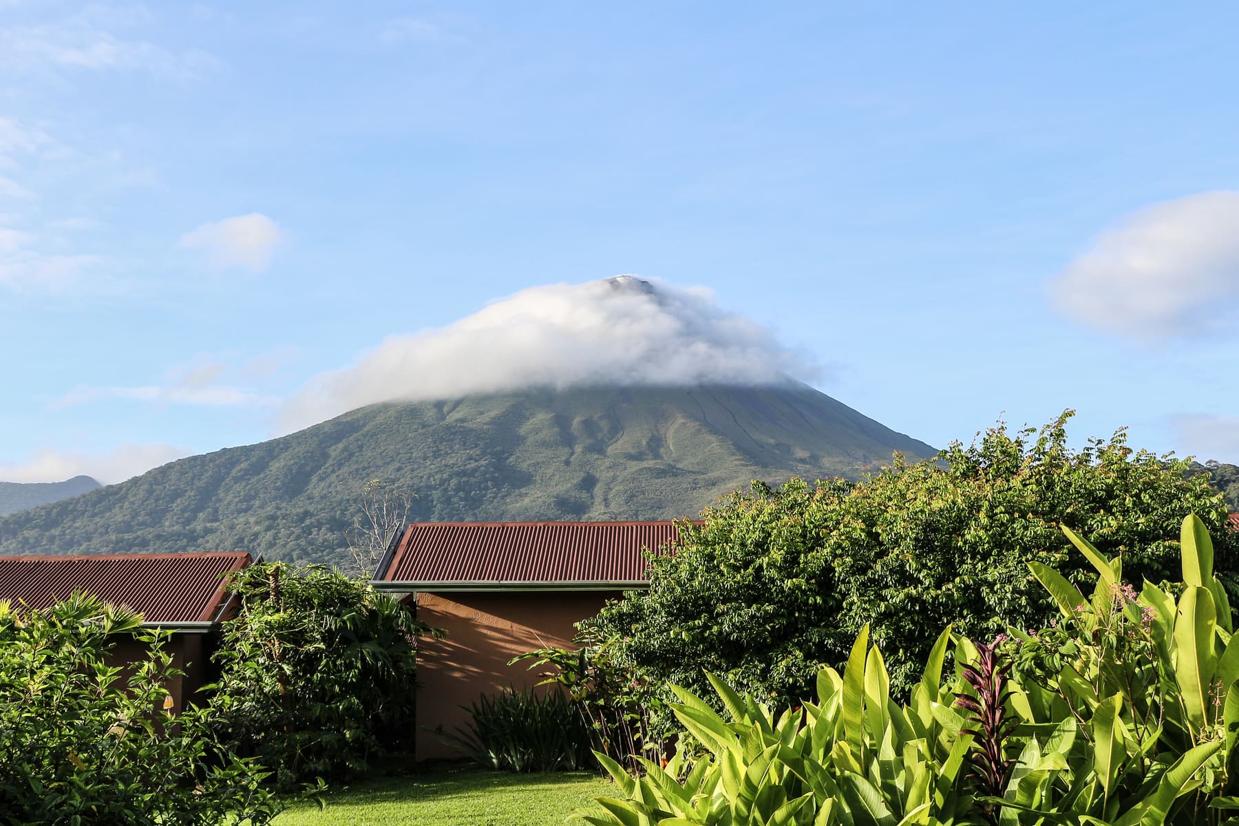 The cloud-capped cone of Arenal Volcano rising above tropical vegetation and low red-roofed buildings