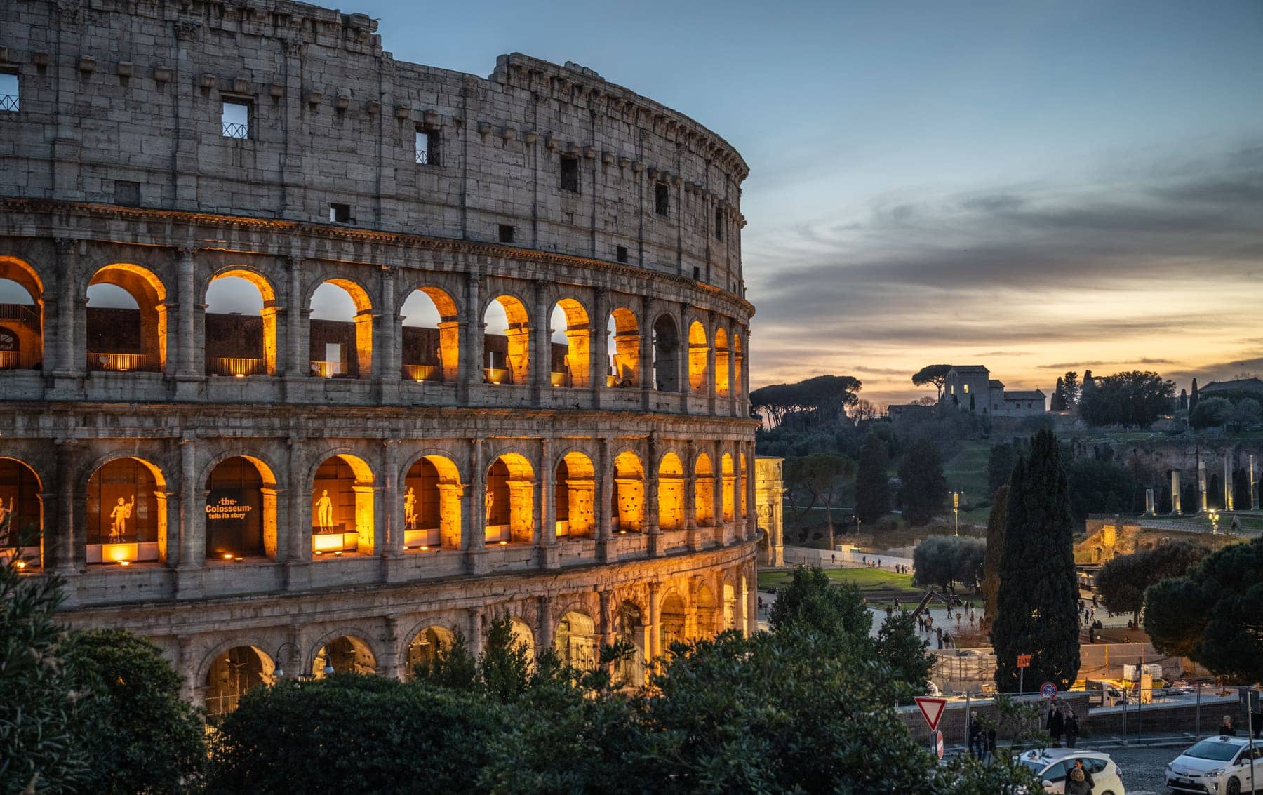 The Colosseum in Rome at dusk with its arches lit, and the Roman Forum visible to the right