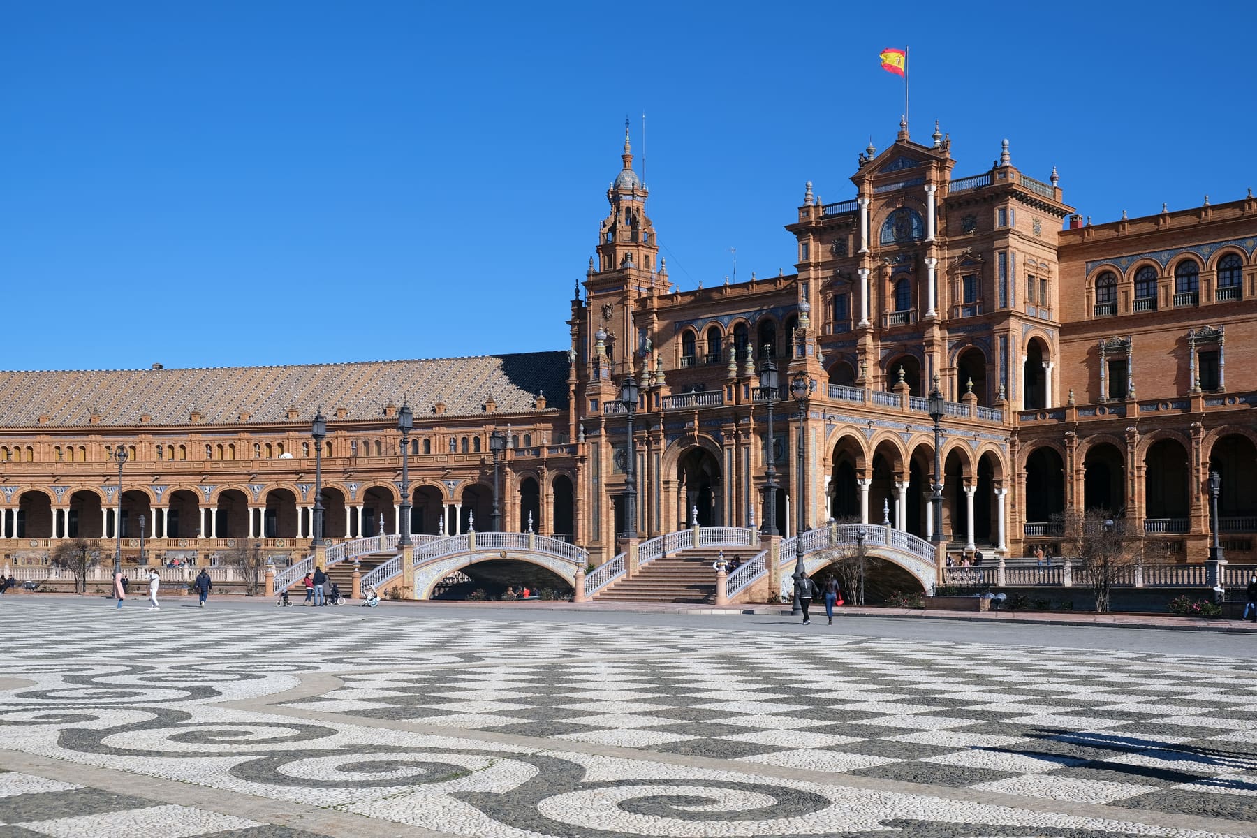 The semicircular brick and tile building of the Plaza de España in Seville with a decorative bridge and patterned paving in the foreground