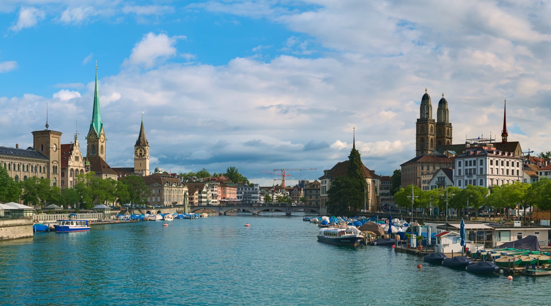 The Limmat river in Zürich with moored boats, the twin towers of the Grossmünster and church spires of the old town under a partly cloudy sky