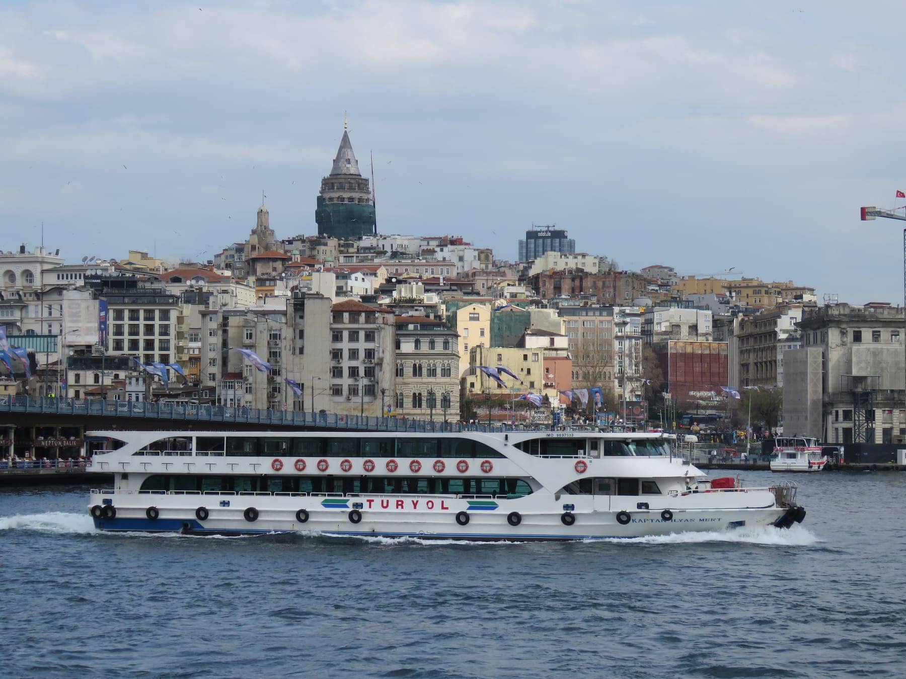 A passenger ferry passing the Galata Bridge, with Galata Tower and hillside buildings behind it in Istanbul