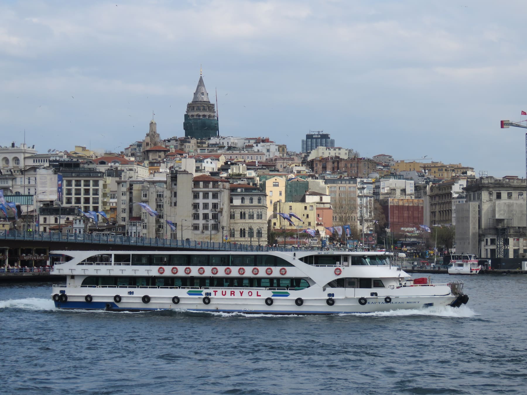 A passenger ferry passing the Galata Bridge, with Galata Tower and hillside buildings behind it in Istanbul